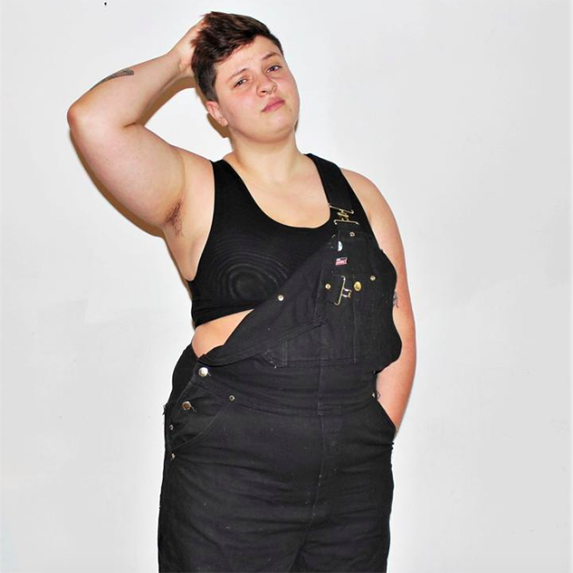 A person with short dark hair wearing a black binder and black overalls