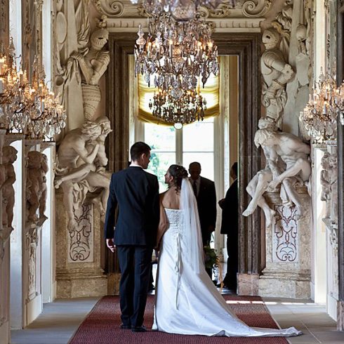 A couple walks down the aisle in a hall with statues and a chandelier