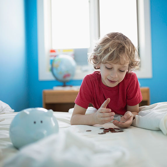 Boy sitting in his blue bedroom with piggy bank on his bed.