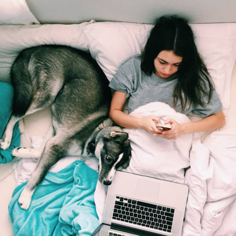 woman in bed with laptop, on the phone, with her pup laying next to her