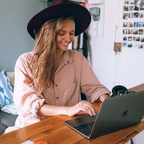 Young woman wearing a hat while smiling and typing on laptop.