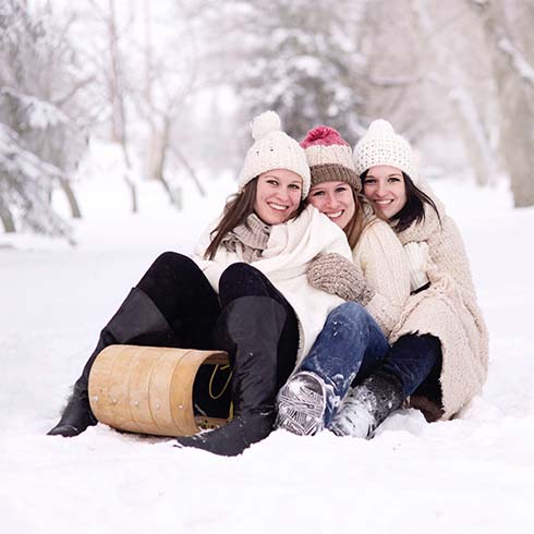 Three young women sit on a tobaggan sled in the snow.