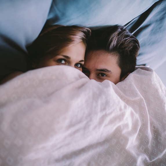 A heterosexual couple peeping over the top of their bedsheet
