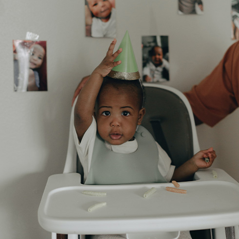 Black baby sitting in a high chair holding a birthday party hat