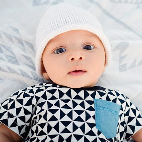 Biracial baby with brown eyes a white hat on his back looking at the camera