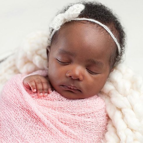 Sleeping baby with curly black hair wearing a white bow, swaddled in a blanket.