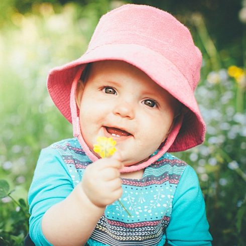 Red-haired baby sitting in a garden with a hat and flower.