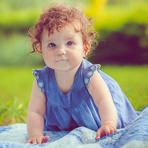 Cute red-haired baby girl with curls and a blue dress on a picnic blanket