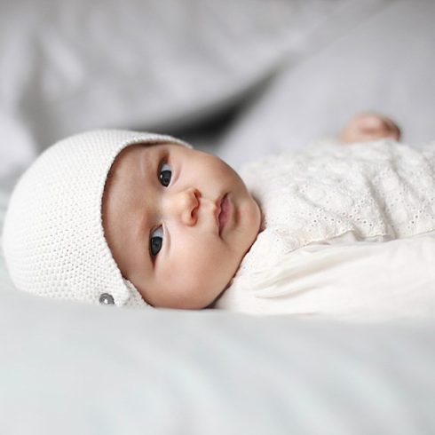 White baby laying on her back in a white hat and white outfit.