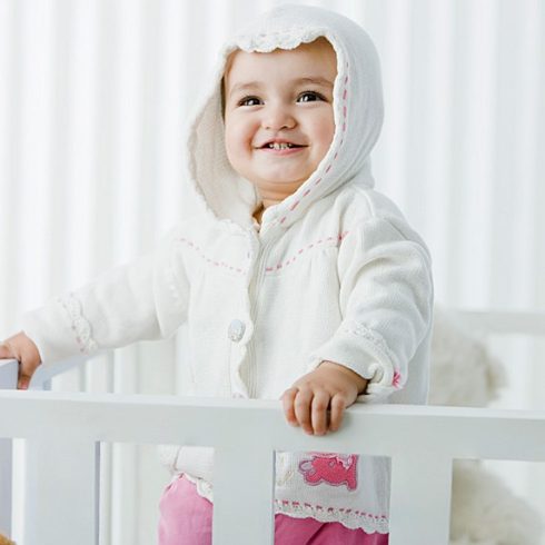 Baby in crib smiling and looking with a hoodie on.