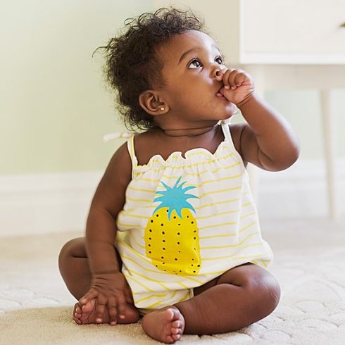 Black baby girl sitting in a pineapple tanktop and sucking her thumb looking off to the side