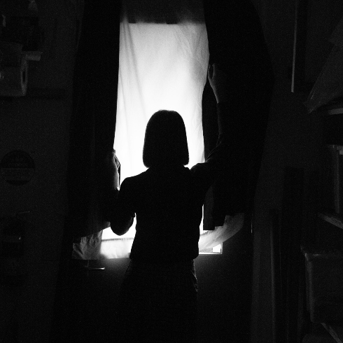 A black and white photo of a woman standing in front of a window in a dark room.