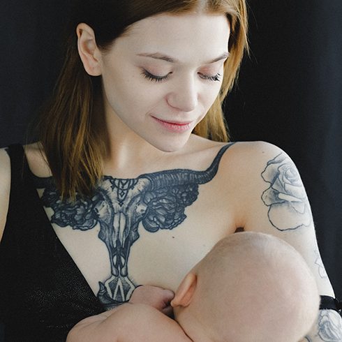 Young woman with tattoos on her chest and arms breastfeeding infant.