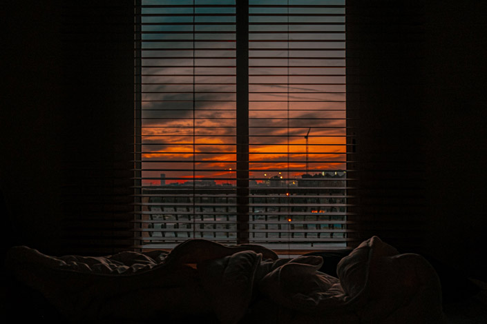 Crumpled blanket at the end of the bed with a view of sunset through a window with blinds
