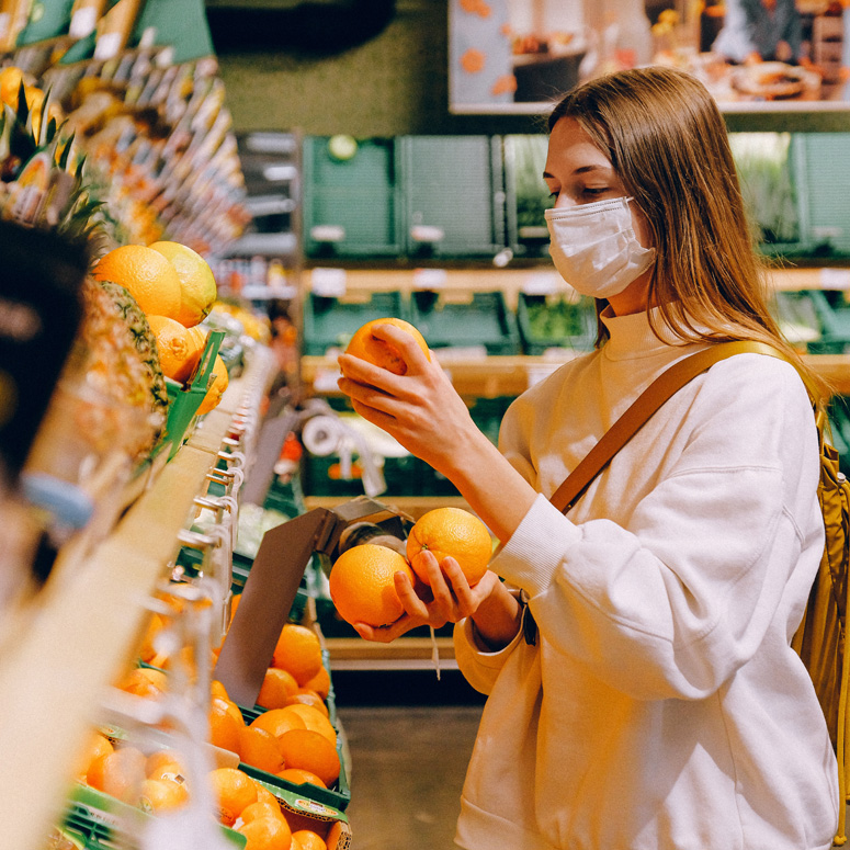 A young woman wearing a face mask picking out oranges at a grocery store.