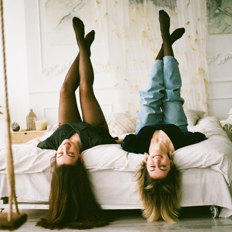 Two women lying on their backs with their legs up in the air, hair hanging off the foot of the bed in a room