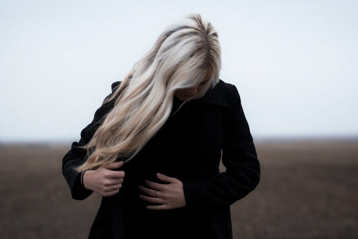 Blonde woman standing in open field looking down at her hand over her stomach