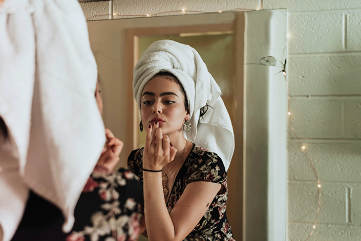 a young white woman with a towel on her head, applying lip gloss while looking in the mirror