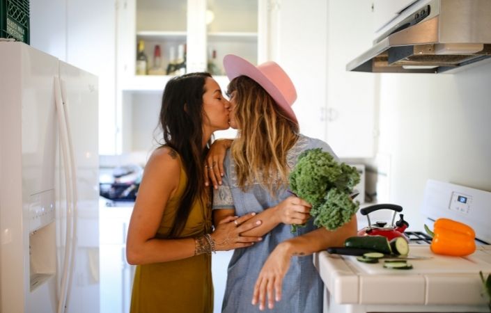 Two women wearing dresses, kissing on the lips in a kitchen while holding a bunch of kale. 