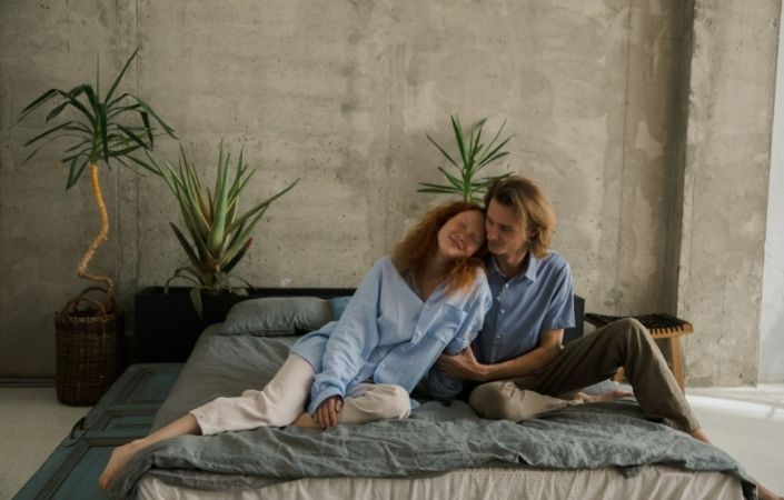 A man and woman in bed, wearing clothes, cuddling and smiling. 