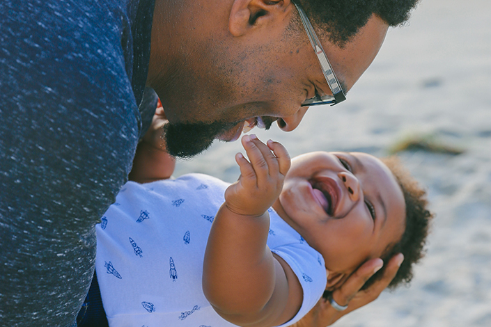 a Black man holds a laughing baby boy in his arms at the beach
