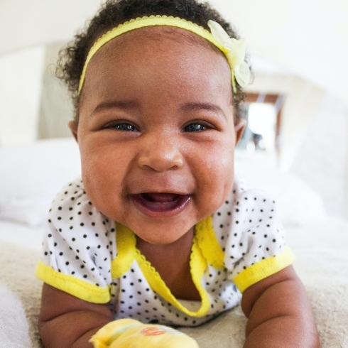 Smiling baby wearing a hair bow and a yellow and gray onesie.