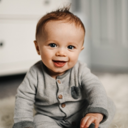 Baby smiling in a gray onesie in his bedroom.