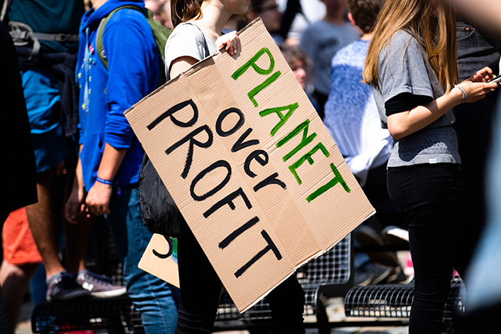 a white woman's hand holding a cardboard sign that reads 'Planet Over Profit'