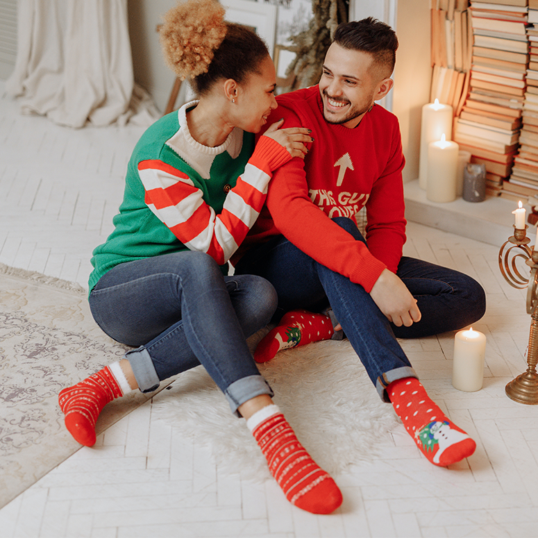 A couple wearing colorful holiday sweaters and socks sits on the floor