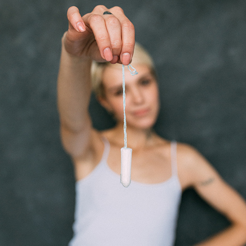 A young Caucasian woman with short blonde hair wearing a white tank top dangling a tampon by its string out in front of her.