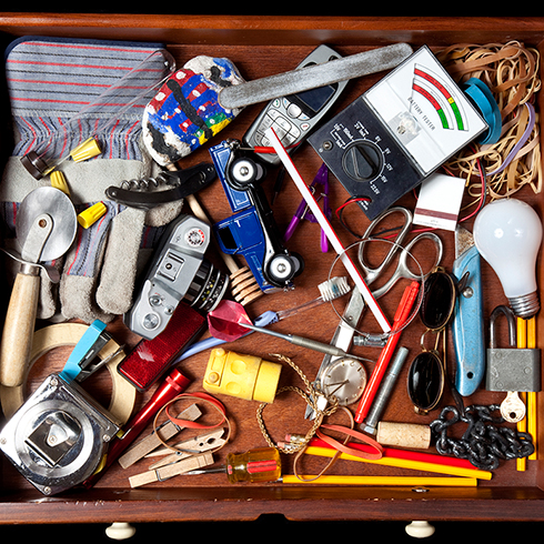 Birds-eye view of a drawer containing a mess of items from gardening gloves to old mobile phones, jewellery, tape and elastic bands.