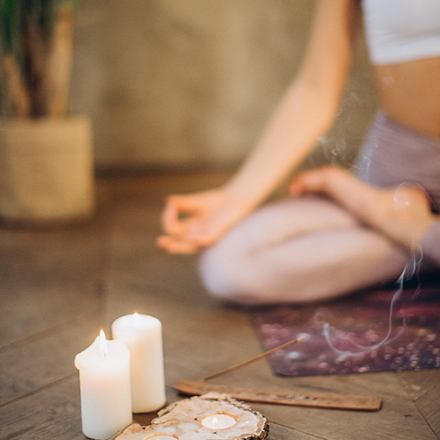 Woman in background meditating on the floor in lotus position, with candle and incense burning in the foreground.