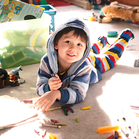 A cute Asian child lying on his stomach with his hoodie hood over his head on a carpeted floor with lots of crayons spread of the floor and toys in the background.