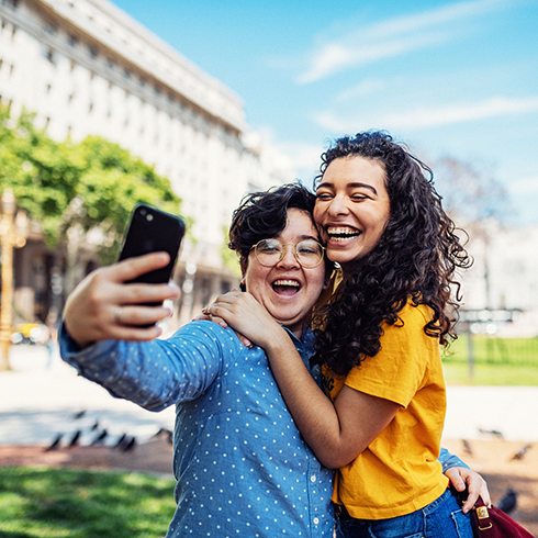 A photo of a happy young lesbian couple taking a selfie outside on a sunny day in front of a buildings and green trees.