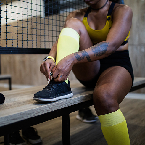 A Black woman, wearing a sports bra, shorts and knee-high yellow socks, sitting on a wooden bench lacing up running shoes in the change room of a gym.