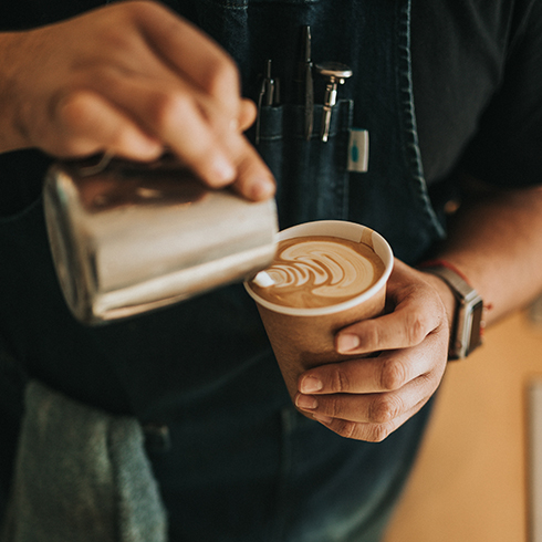 A barista wearing an apron, pouring milk into an espresso coffee in a disposable cup.