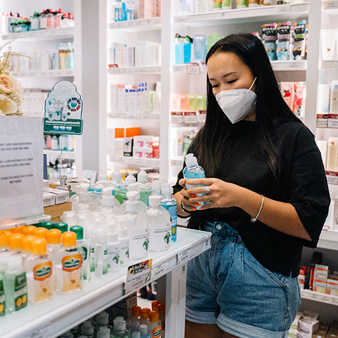 A young Asian woman wearing a black tee shirt, denim shorts and an N95 face mask in a drug store shopping for hand sanitizer and reading the label of a bottle she is holding.