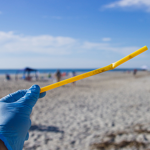 A hand wearing a blue latex glove holding up a bent yellow plastic straw with an out of focus beach in the background.