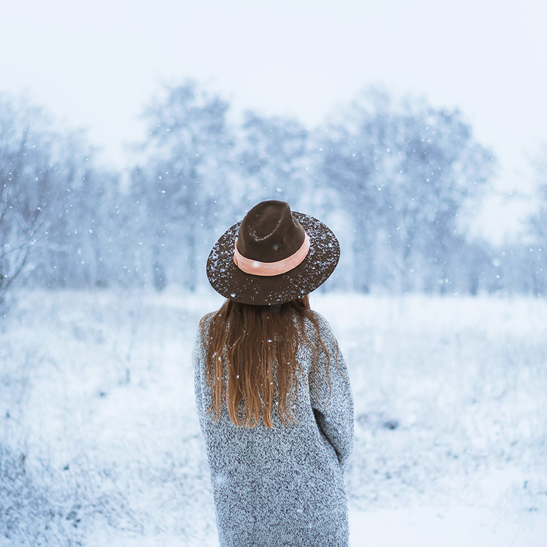 a young woman with long hair and a hat standing in the snow with her back to the camera