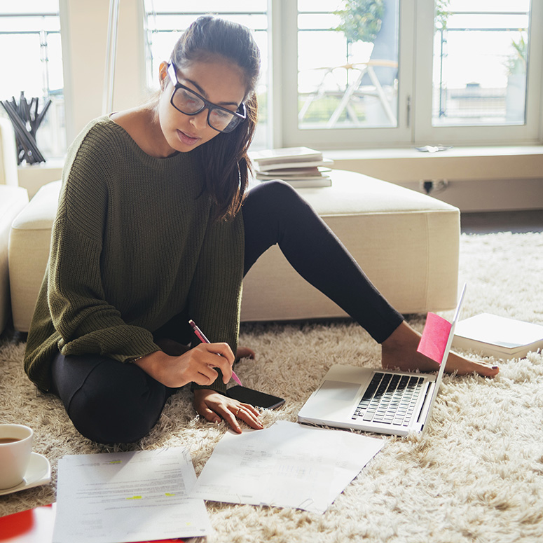 a young woman with glasses sits on the floor of her condo looking at her finances with an open laptop
