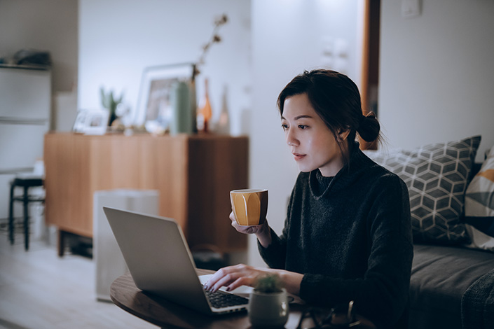 a young Asian woman sitting coffee from a mug in her apartment while looking at her open laptop