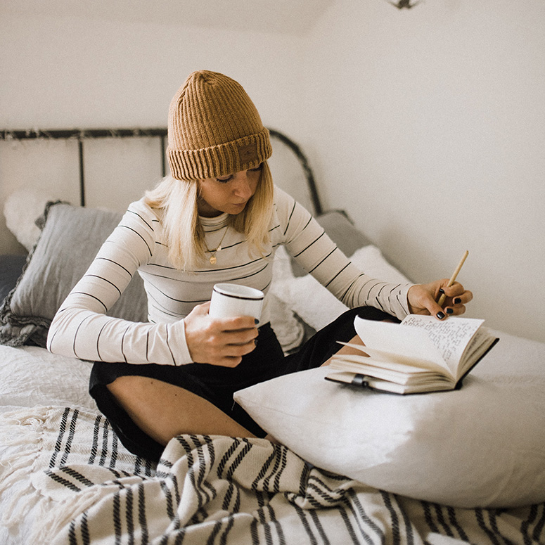 Woman sitting on her bed holding a mug and writing in a notebook