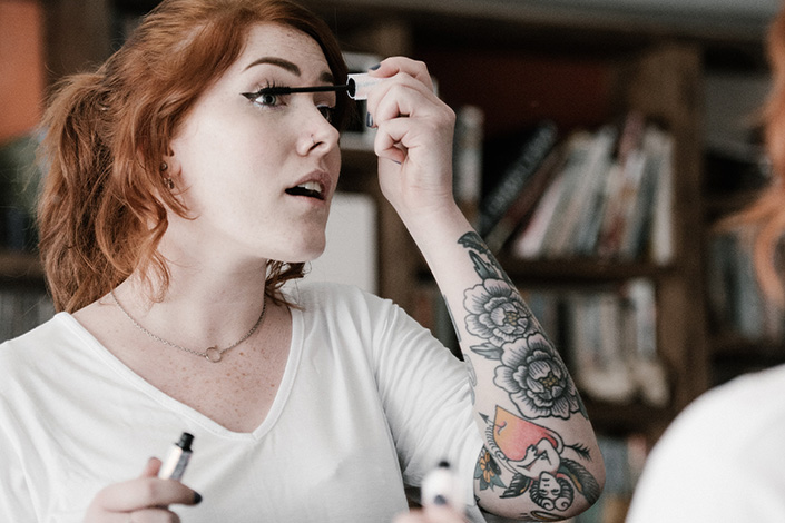 a young white woman with tattoos applies eye makeup in the mirror