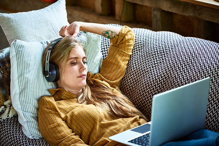 a white woman in a yellow sweater lying on a couch with headphones, looking at her open laptop