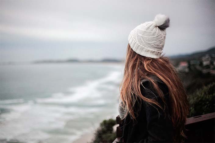 woman with long brown hair and a white hat looking at ocean