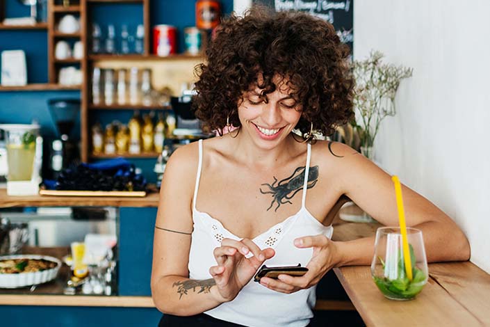 A young woman with tattoos is taking a break in a colourful cafe, browsing the internet on her smartphone.