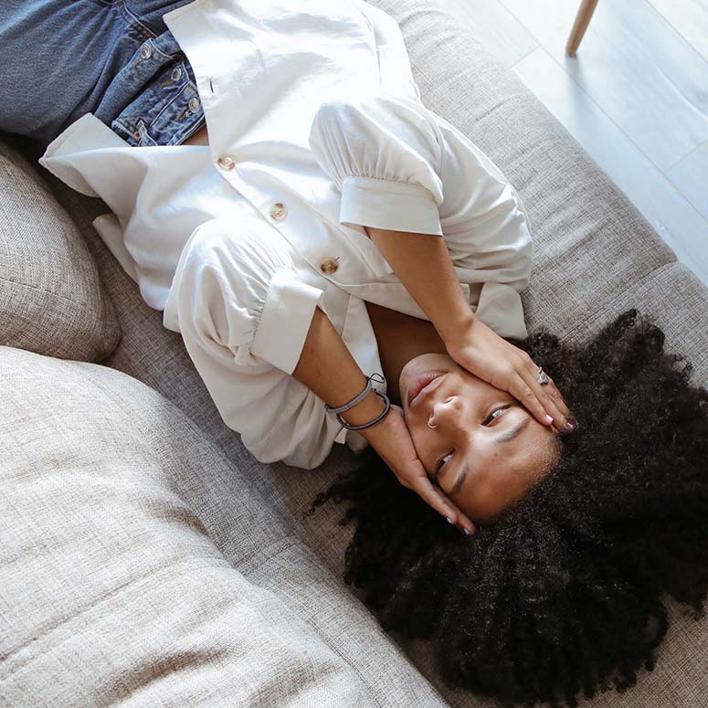 Black woman wearing a white shirt lays on couch with her hands on her head