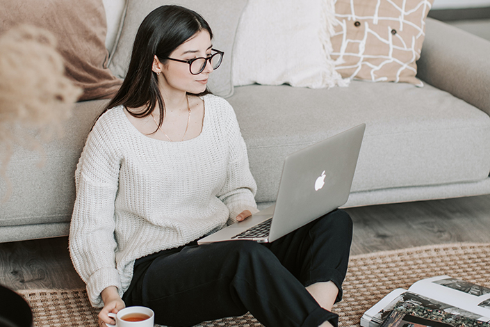 a young white woman with glasses sitting on the floor of her apartment with her laptop and a cup of coffee