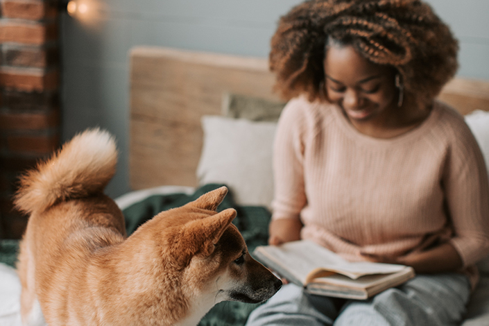 a young Black woman smiles while in bed reading, with her dog at her feet