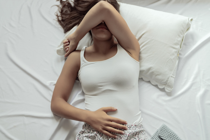 Young woman in discomfort lies in bed with one arm laid over her face